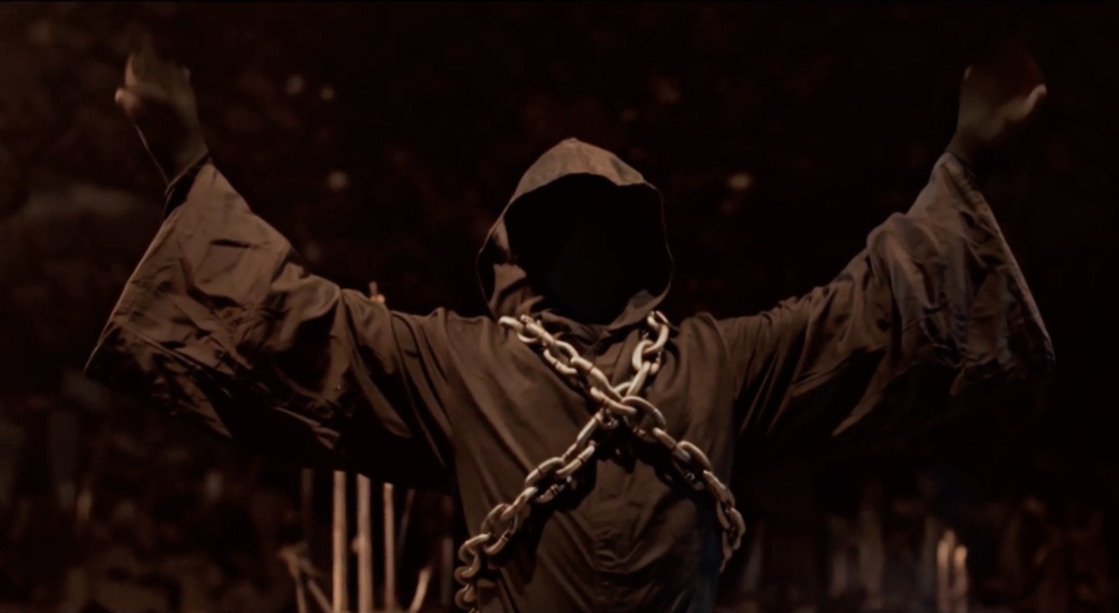 A hooded figure wrapped in chains raises both arms against firelight